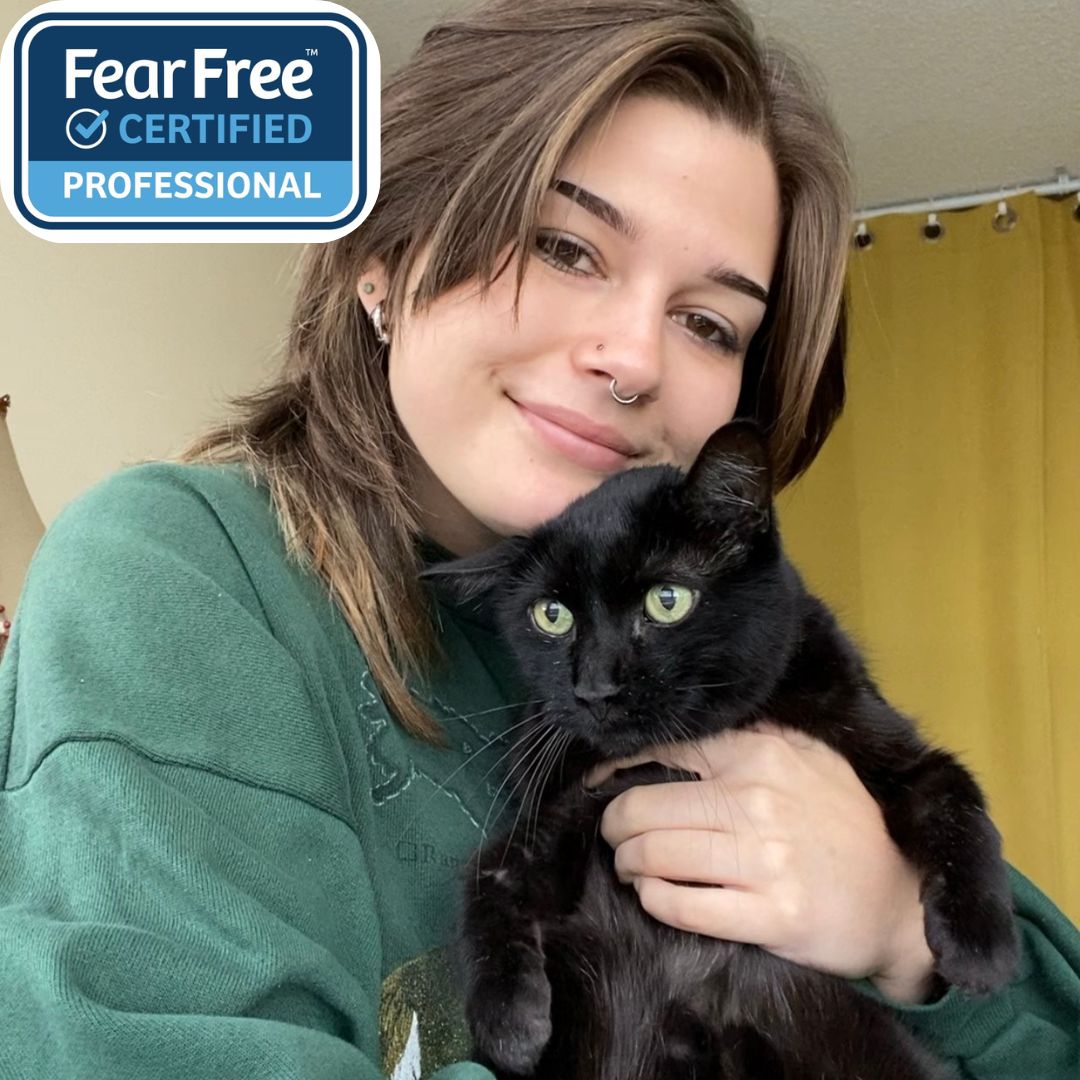 Tania, a Veterinary Technician at Waterloo West Animal Hospital, smiles warmly while holding a sleek black cat in her arms. She wears a green sweatshirt and a nose ring, with a “Fear Free Certified Professional” badge in the corner, emphasizing her gentle, cat-friendly care approach in a cozy indoor setting.